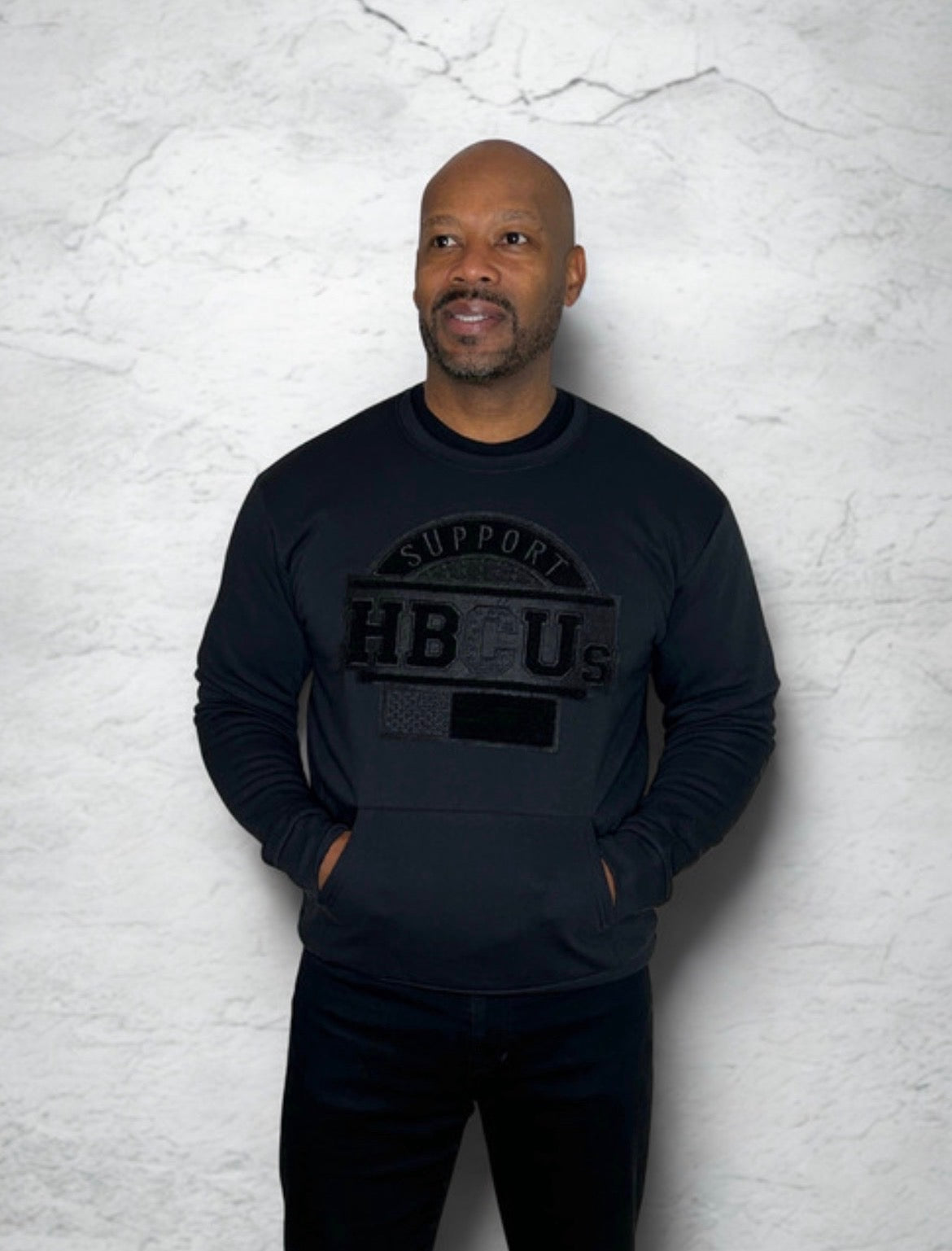 Man wearing an all-black SUPPORT HBCUs sweatshirt featuring a hand-stitched chenille patch and front pockets, representing support for Historically Black Colleges and Universities