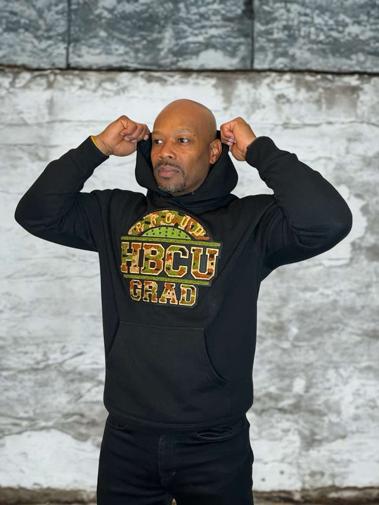 Model wearing a black Proud HBCU Grad hoodie with a handcrafted camo chenille patch, varsity-style stitched lettering.